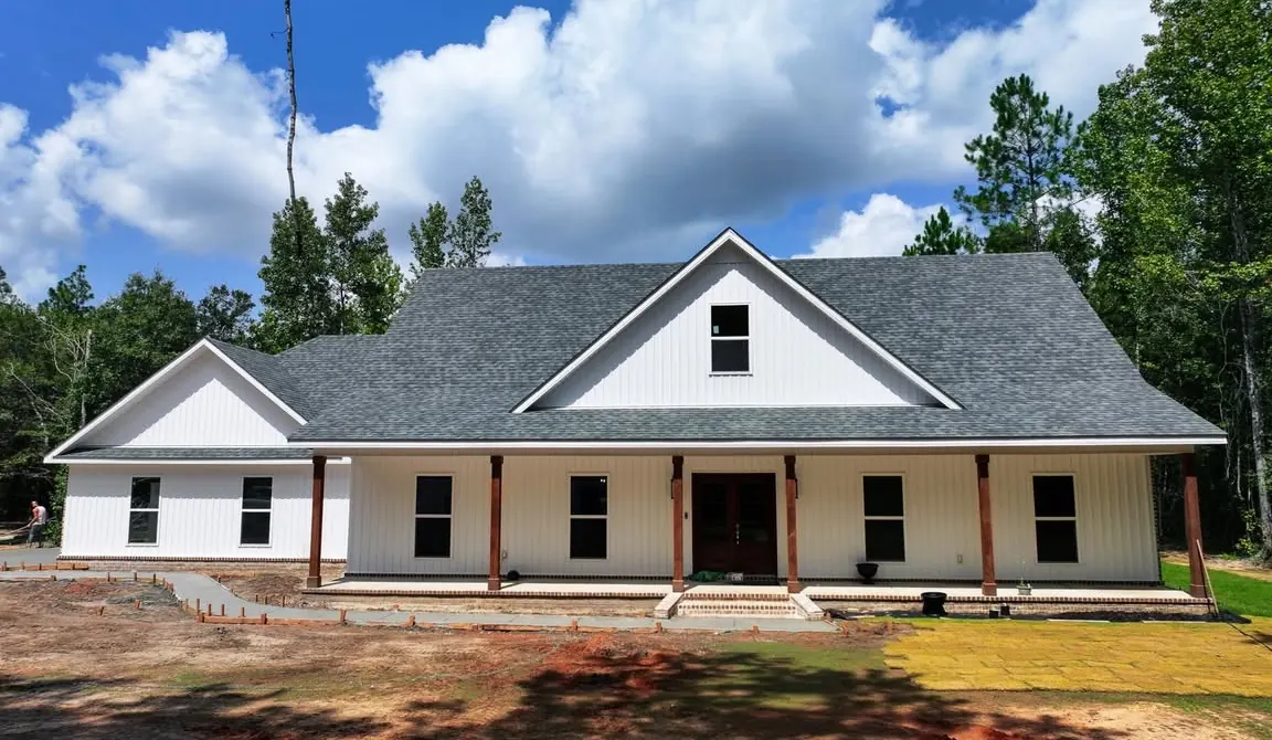 Aerial view of a new white farmhouse with a long front porch and cedar posts, built by Byrd, surrounded by pine trees